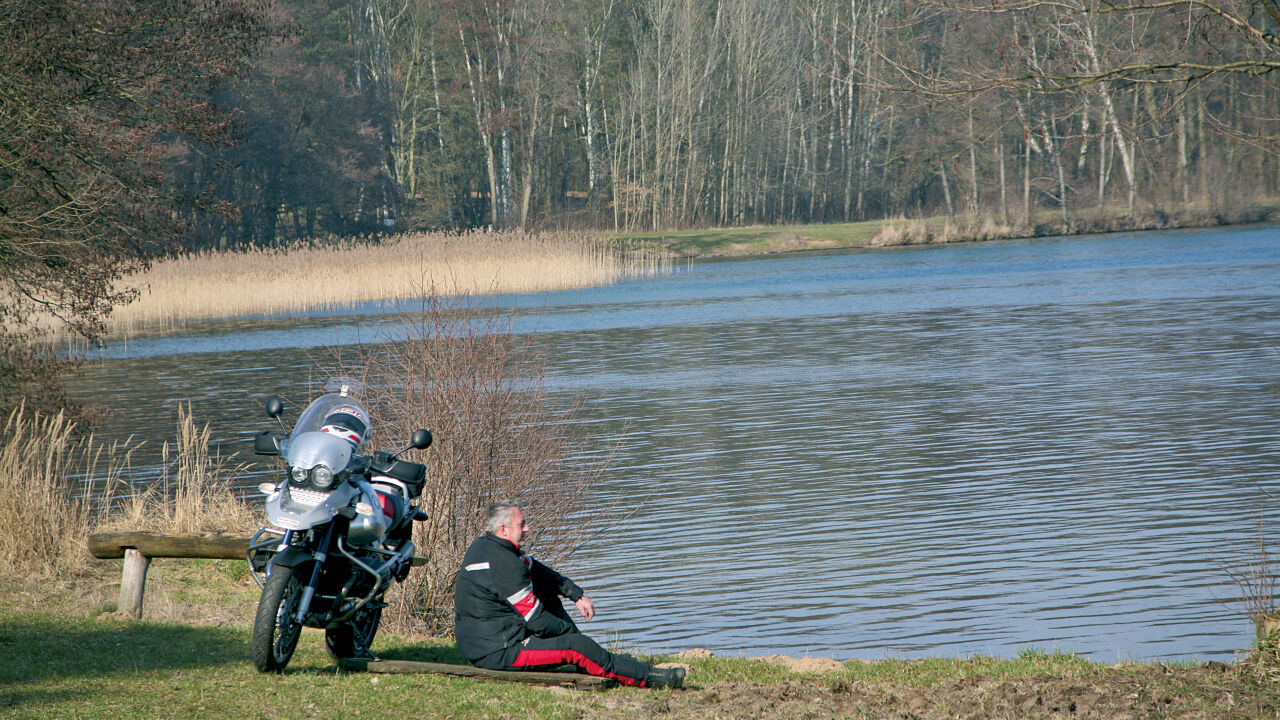 Motorradfahrer sitzt am Ufer neben seiner Maschine, vor ihm ein See mit Schilf Motorradfahrer sitzt am Ufer neben seiner Maschine, vor ihm ein See mit Schilf